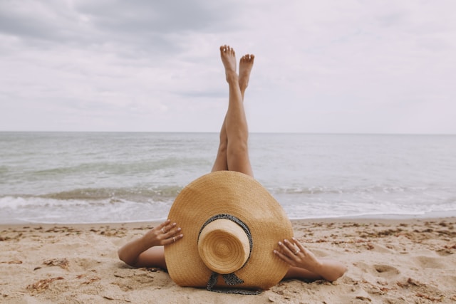 Woman laying on the beach with her legs up in the air. She is wearing a straw hat.